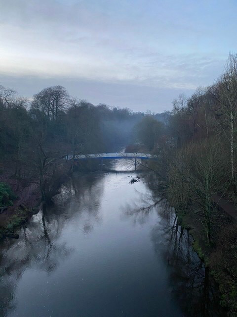 autumn evening. treeless canopies border the river kelvin. the humpback bridge is in the distance. fot sits over the water and trees. the sky is skiffed with thin clouds.